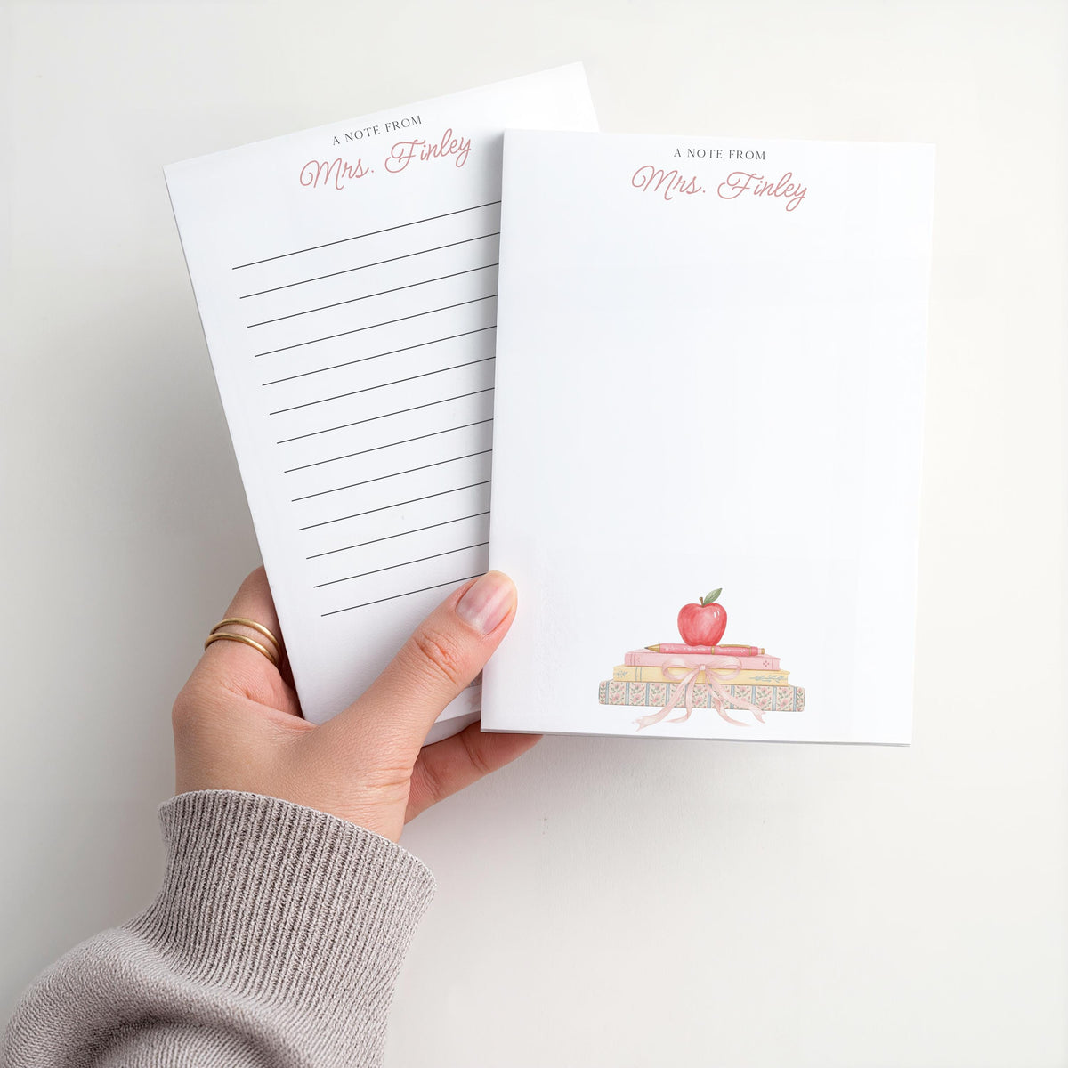 May include: White notepad with the text "A Note From Mrs. Finley" in pink script. The notepad features a watercolor illustration of a stack of books topped with a red apple. One notepad has lined pages.